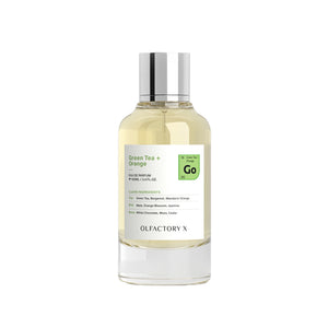 A clear glass bottle of Olfactory X Green Tea + Orange Eau de Parfum with a silver cap, containing a pale yellow liquid, set against a plain white background.