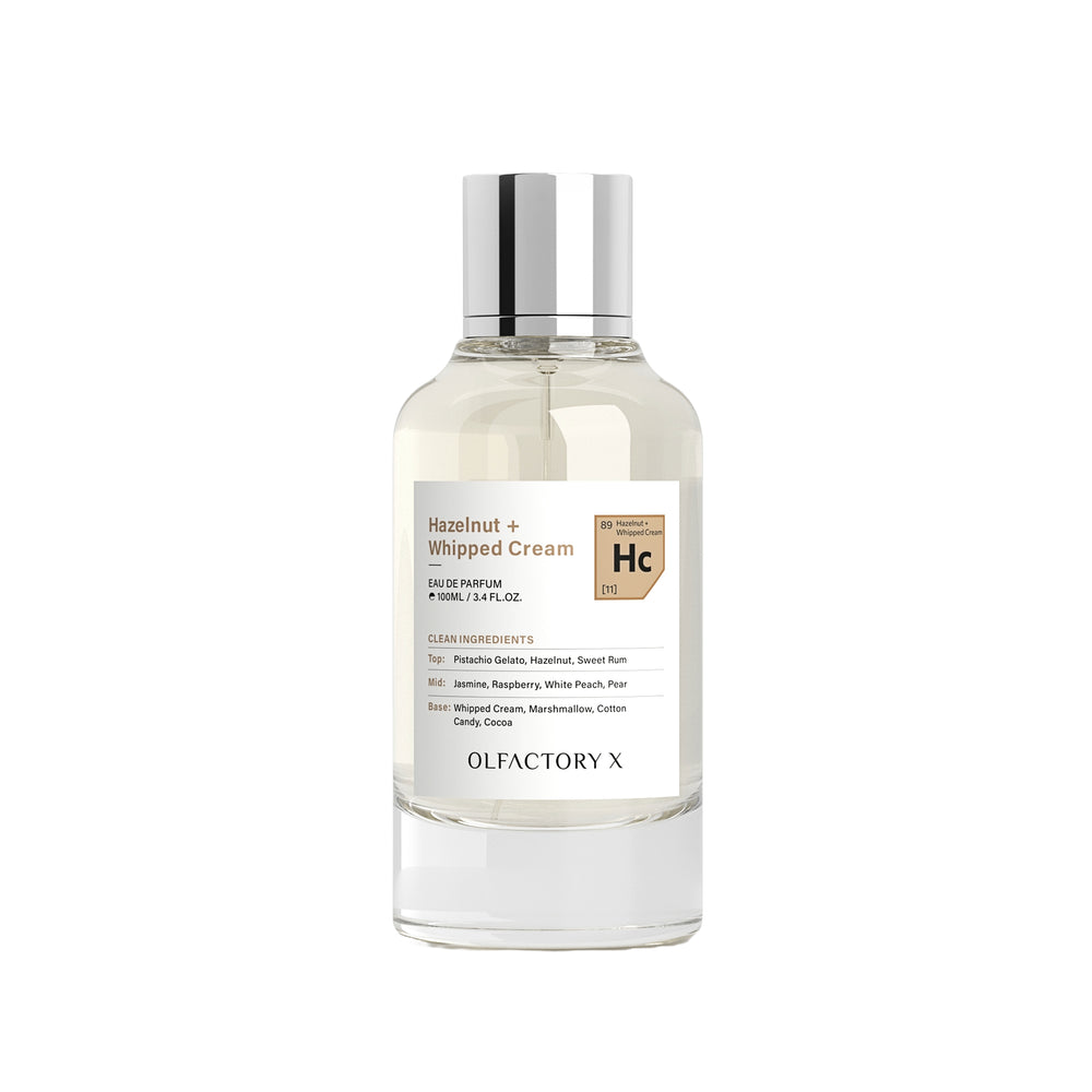 A clear glass bottle of Olfactory X Hazelnut + Whipped Cream perfume with a silver cap, shown on a plain white background.