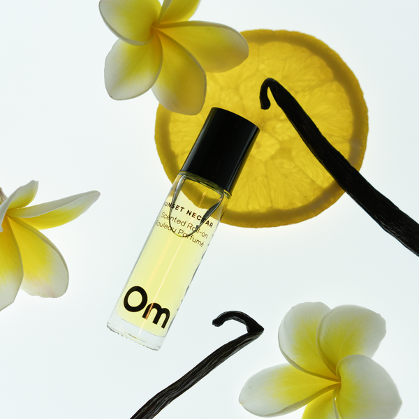 OM ORGANICS Sunset Nectar Scented Roll On surrounded by plumeria flowers, vanilla bean stocks and lemon slice