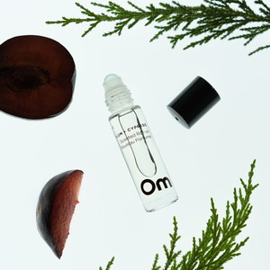 OM Organics Wild Plum and Cypress scented roll-on perfume surrounded with cypress leaves and plums on off white background