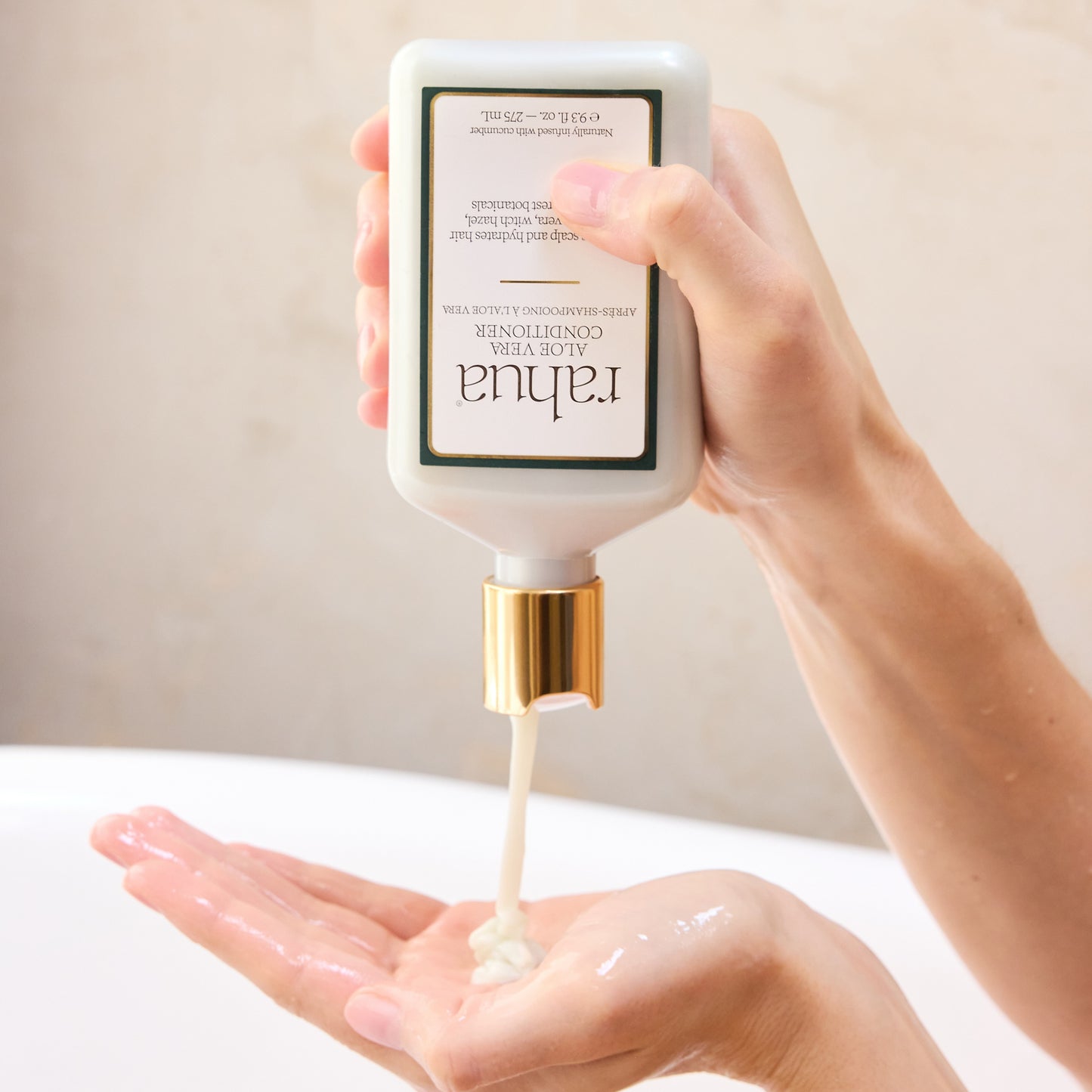 Hand holding a bottle of skincare product labeled 'Rahua Aloe Vera Conditioner' with cream being dispensed onto a palm.