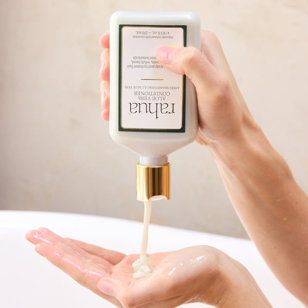 Hand holding a bottle of skincare product labeled 'Rahua Aloe Vera Conditioner' with cream being dispensed onto a palm.