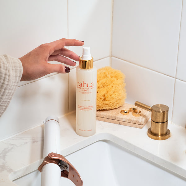 A woman is reaching to grab a bottle of Rahua Heat Protectant Shield in a bathroom setting.