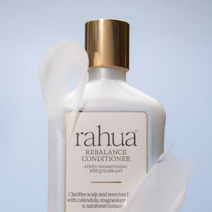 A bottle of Rahua Rebalance Conditioner with a gold cap, shown against a light blue background with white conditioner cream splashed around it.