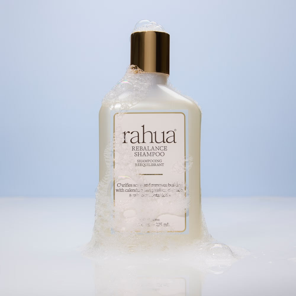 A bottle of Rahua Rebalance Shampoo with a frosted off-white body, a metallic gold cap, and a cream-colored label with a light blue border, set against a plain white background.
