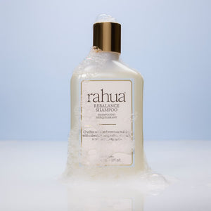 A bottle of Rahua Rebalance Shampoo with a white body, gold cap, and a clear gel-like substance dripping from the bottom, set against a light blue gradient background.