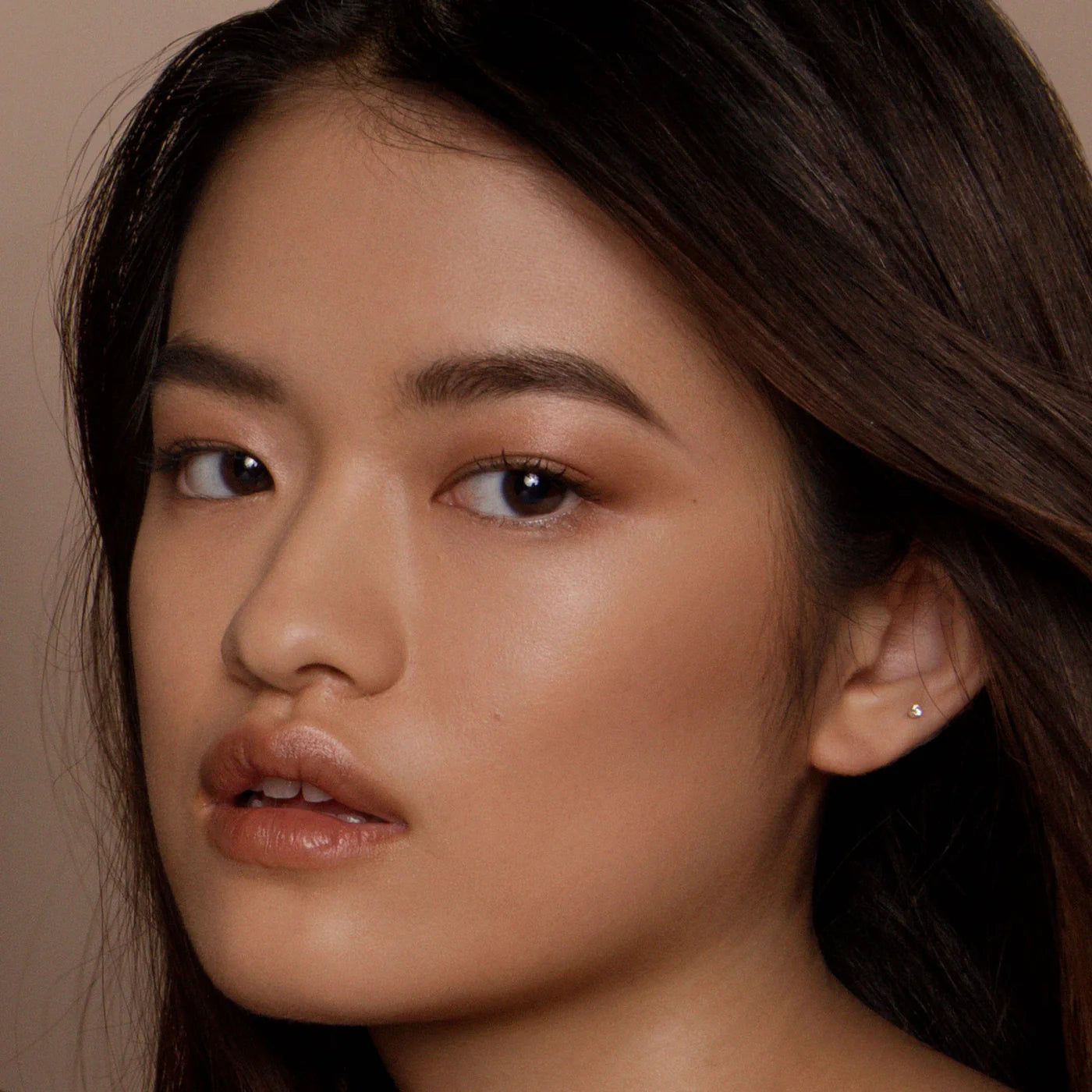 Close-up of a person with dark hair and neutral makeup against a beige background. RITUEL DE FILLE Inner Glow Crème Pigment Eros