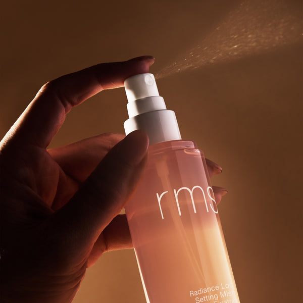 Hand holding a RMS beauty product with a warm, brownish background. RMS BEAUTY Radiance Lock Setting Mist