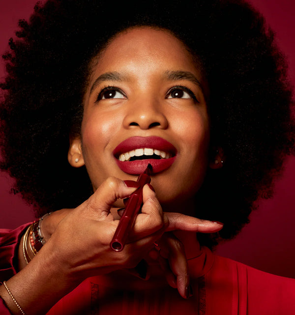 Woman applying red lipstick with Ruby Hammer lip liner brush against a red background
