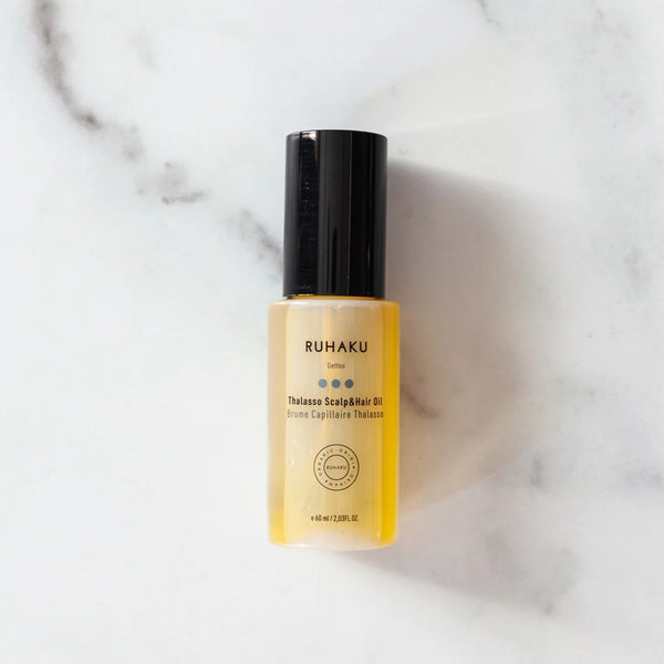 A bottle of Ruhaku Thalasso Scalp Hair Oil with a black cap and yellow liquid inside, placed on a white marble surface.