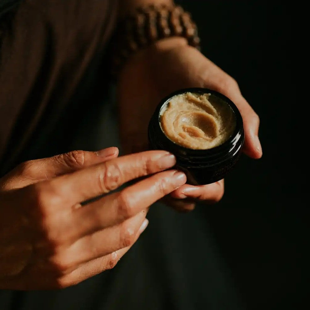 Person holding a black jar of beige colored SIAM SEAS MEKHA Exfoliating Cleanser against a dark background