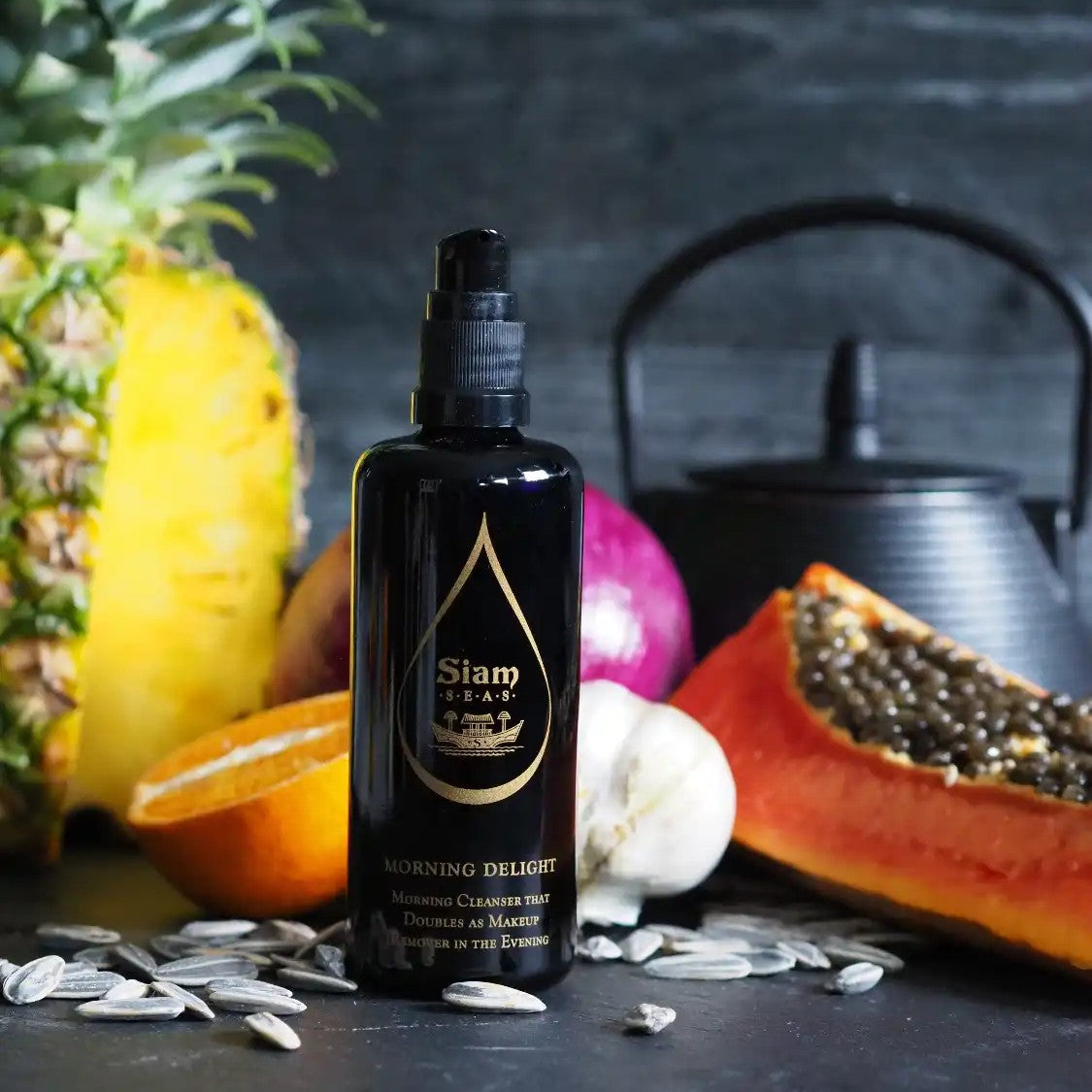 Black bottle of 'SIAM SEAS Morning Delight Cleanser and Makeup Remover' product with fruits and a teapot in the background