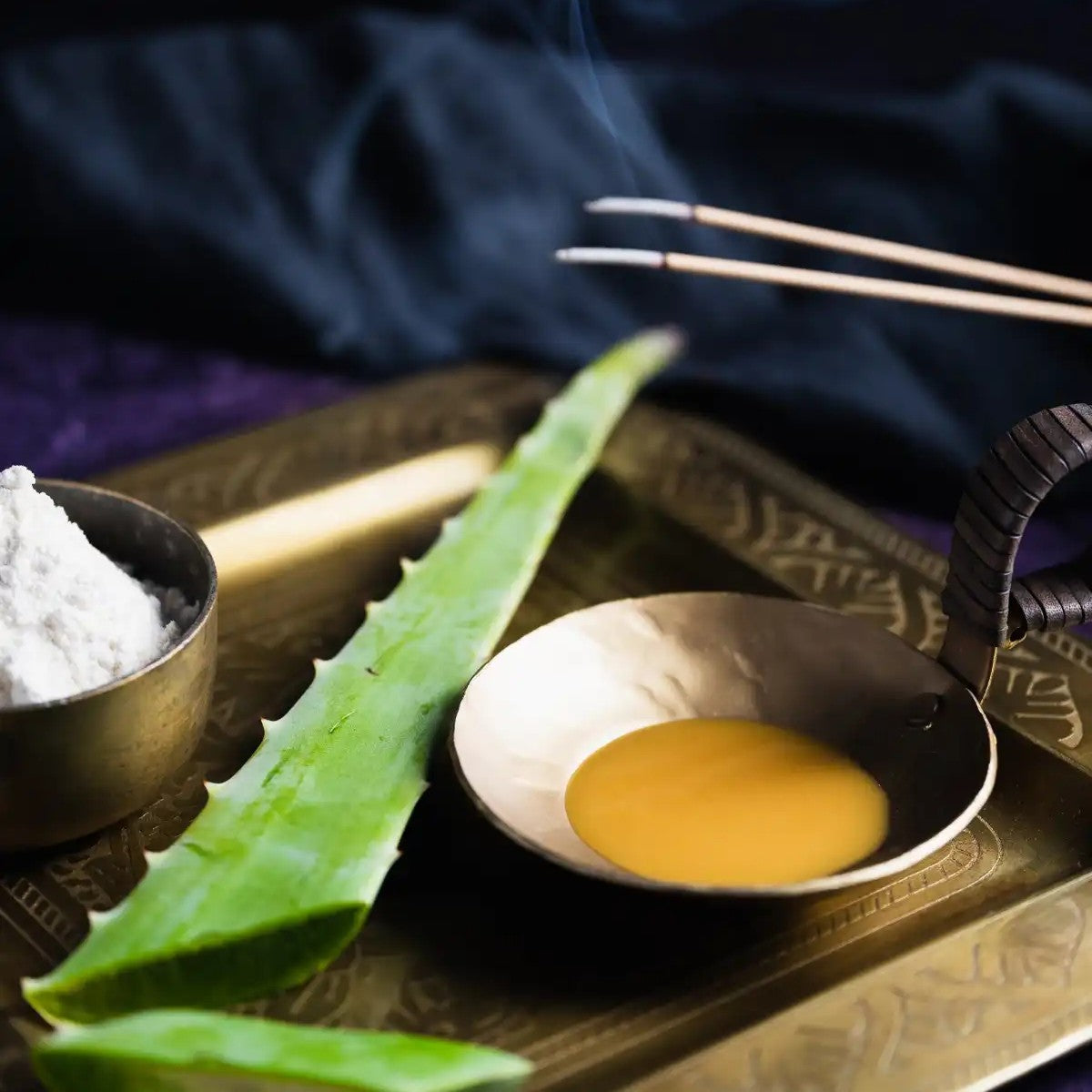 Aloe vera leaf, a bowl of yellow liquid, and a small container of white powder on a decorative tray with incense sticks.