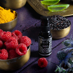 Bottle with pump of Siam Seas Sai Clear Skin Vitamin C Serum with bowls of raspberries, black rice, and yellow powder on a purple surface.