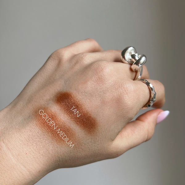 Hand with makeup swatches labeled 'Tan' and 'Golden Medium' on a neutral background