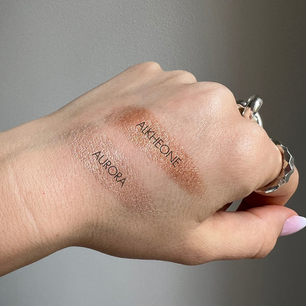 Hand swatching makeup products labeled 'Aurora' and 'Alkheone' on a neutral background
