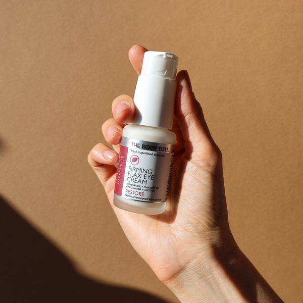 Hand holding a bottle of 'The Body Deli' firming eye cream against a light brown background. 
