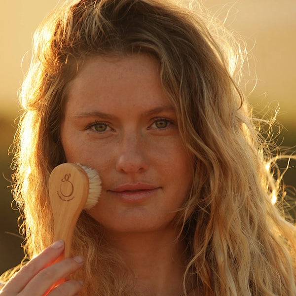 A close-up lifestyle shot of a woman with long blonde hair holding a wooden facial dry brush against her cheek, set against a warm, golden outdoor background.