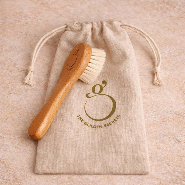 A wooden face brush with cream-colored bristles rests on a beige drawstring pouch printed with the logo and name 'THE GOLDEN SECRETS'. The pouch is placed on a textured beige stone surface.