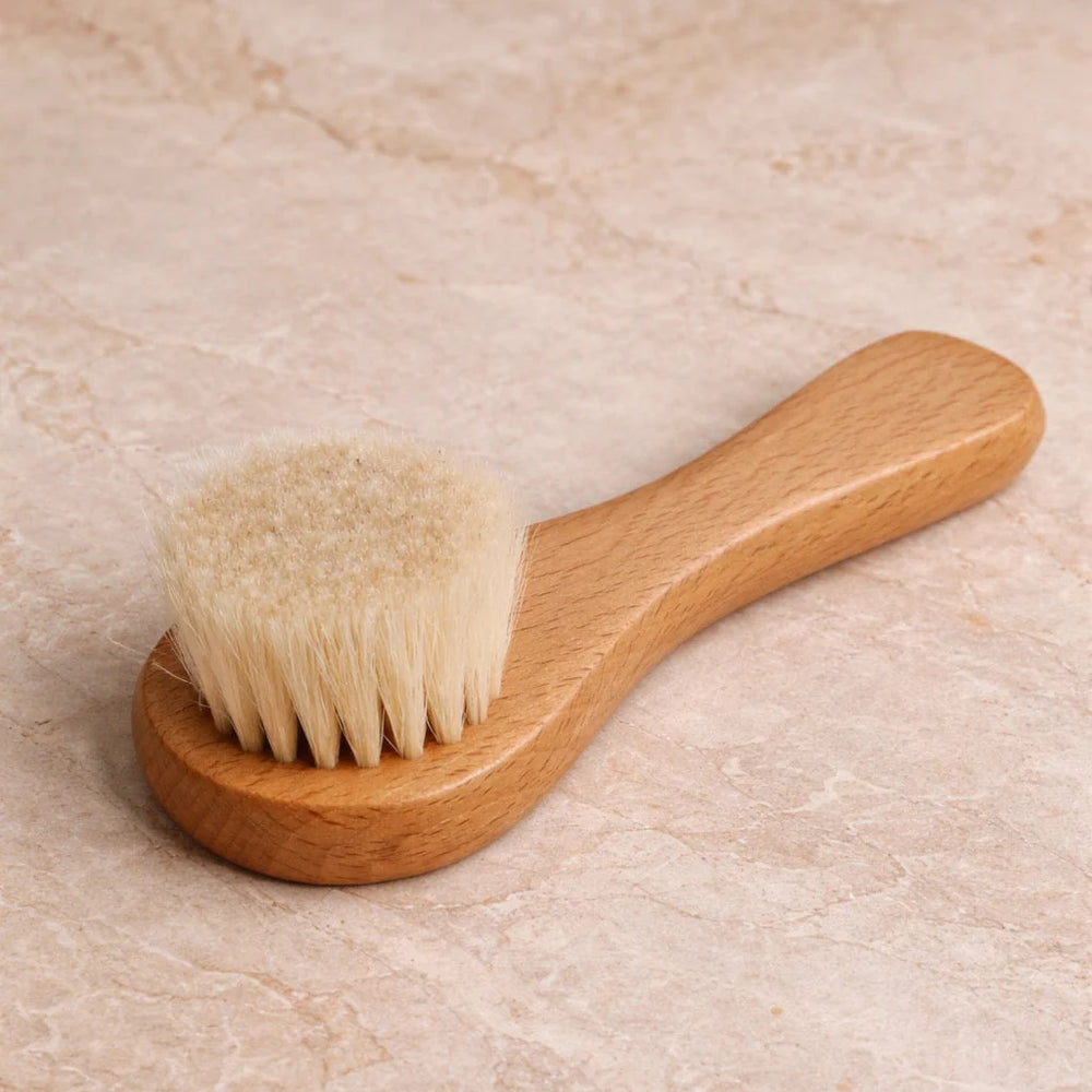 A small wooden face brush with a light natural wood handle and soft cream-colored bristles, shown on a plain white background.