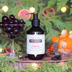 The Potion Cosmetics Embers Body Potion bottle with warm festive background