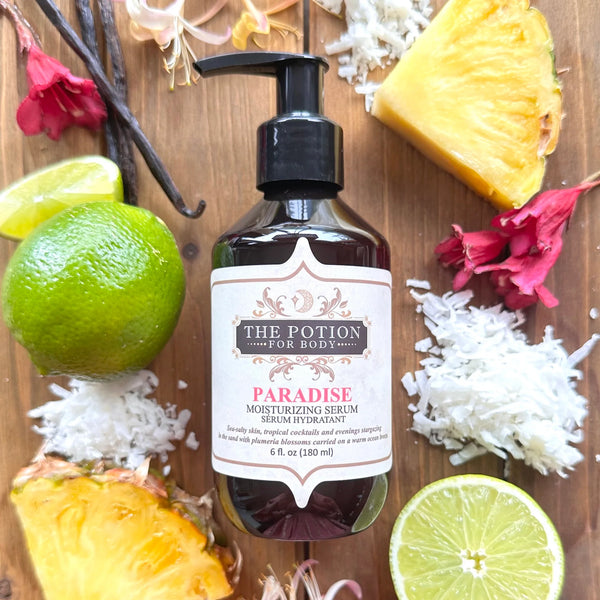 Bottle of 'THE POTION COSMETICS Paradise Body Potion' moisturizing serum surrounded by tropical fruits and flowers on a wooden surface
