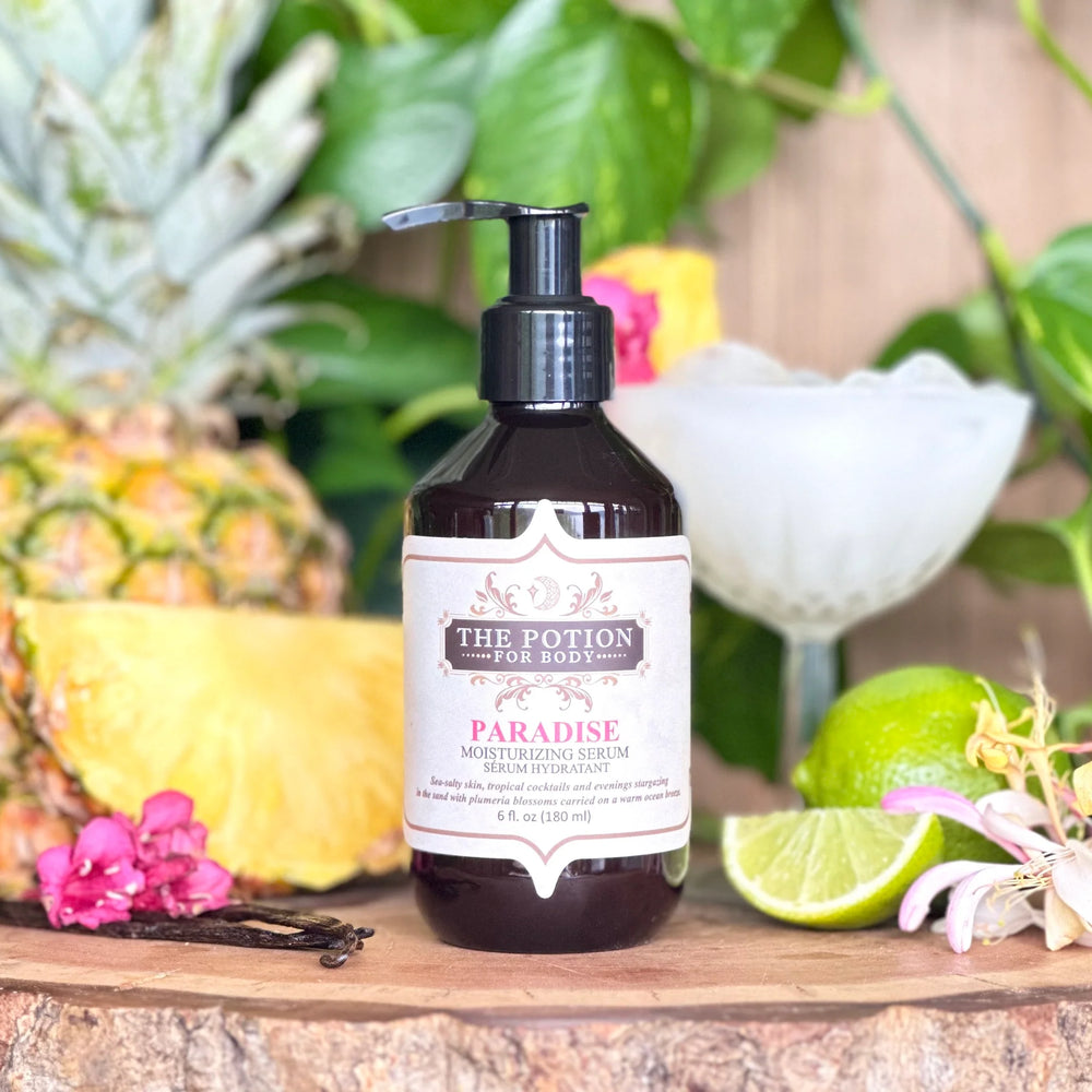 Bottle of 'THE POTION COSMETICS Paradise Body Potion' with a pineapple, limes, and flowers on a wooden surface.
