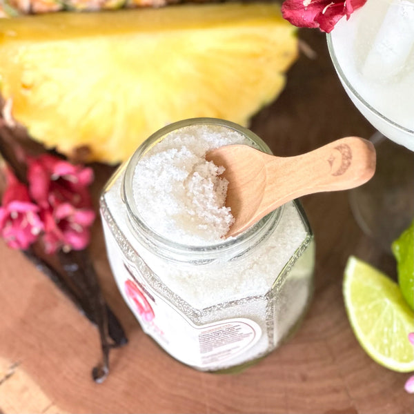 Jar of THE POTION COSMETICS Paradise Mineral Body Scrub with a wooden spoon, vanilla bean, and flowers on a wooden surface
