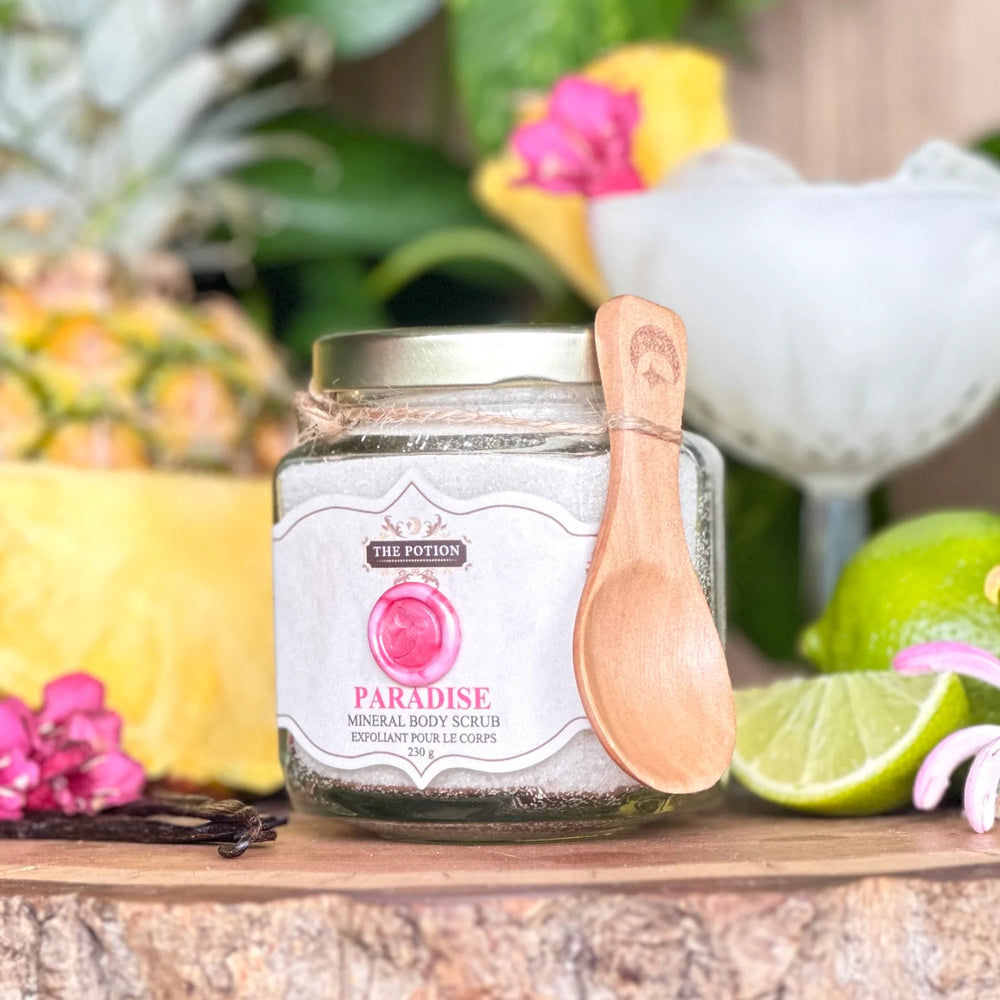 Jar of 'Paradise' mineral body scrub from The Potion Cosmetics with a wooden spoon on a wooden surface with tropical fruits and flowers.
