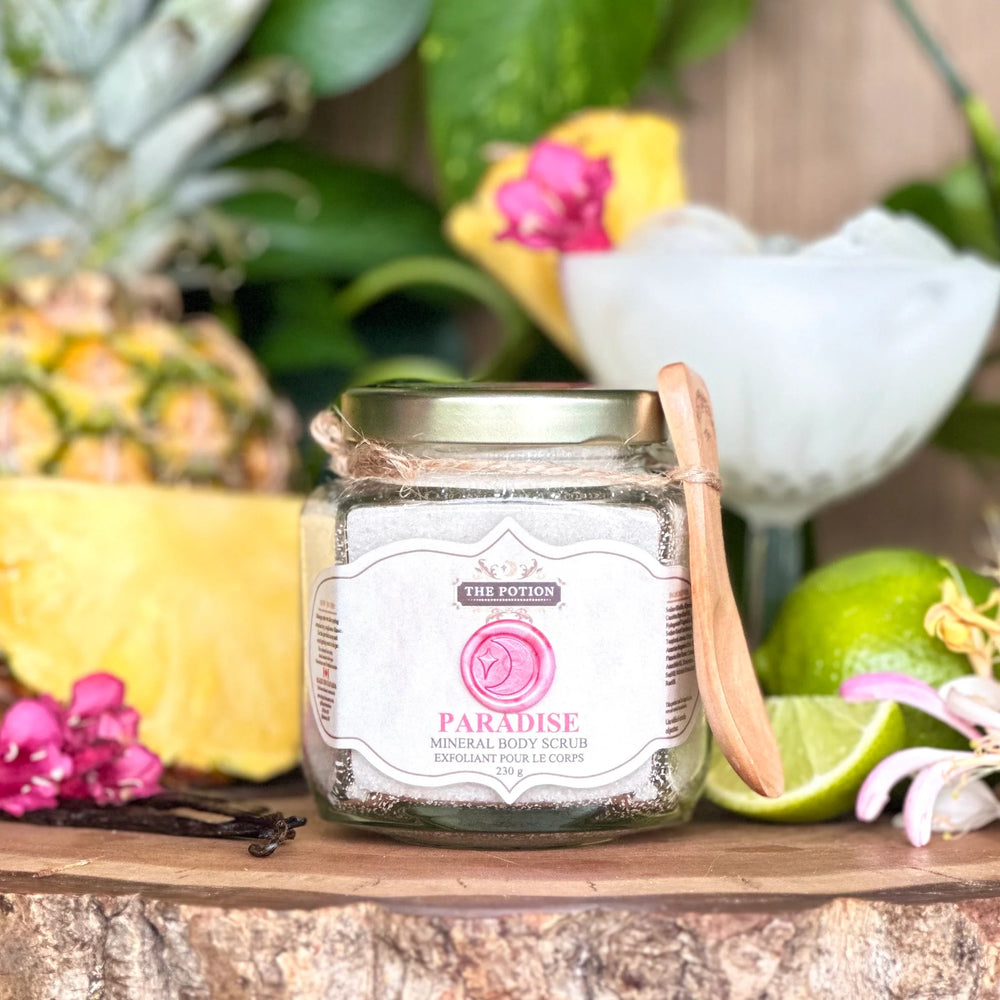 Jar of 'Paradise' mineral body scrub from The Potion Cosmetics with a wooden spoon on a wooden surface with tropical fruits and flowers.