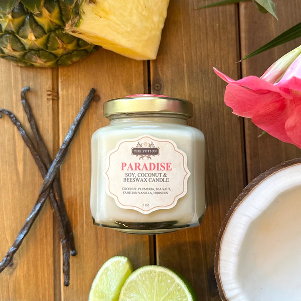 Candle labeled 'Paradise' surrounded by pineapple, vanilla beans, and coconut on a wooden surface.