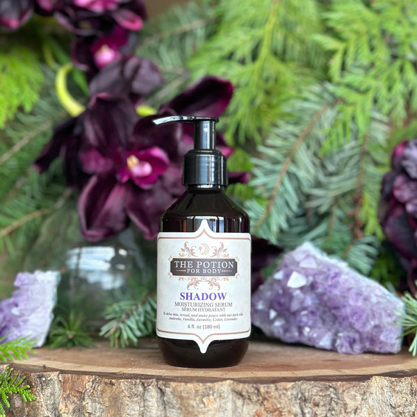 THE POTION COSMETICS Shadow Body Potion bottle surrounded by cedar, jasmine, and vanilla, evoking a deep, sultry floral musk fragrance.