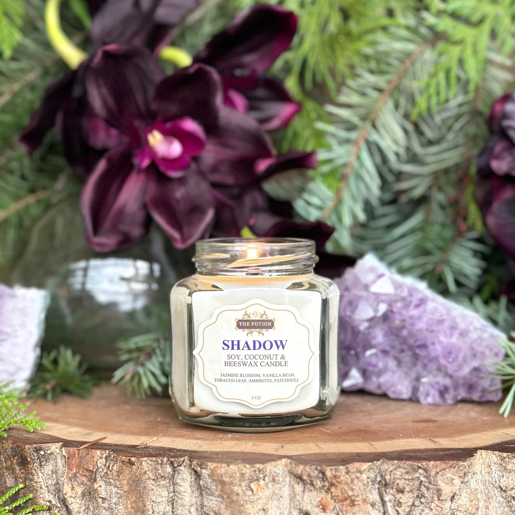 THE POTION COSMETICS Shadow Candle in a glass jar with wooden wick, surrounded by vanilla, jasmine, tobacco leaf, and patchouli, evoking a cozy, uplifting floral aroma.