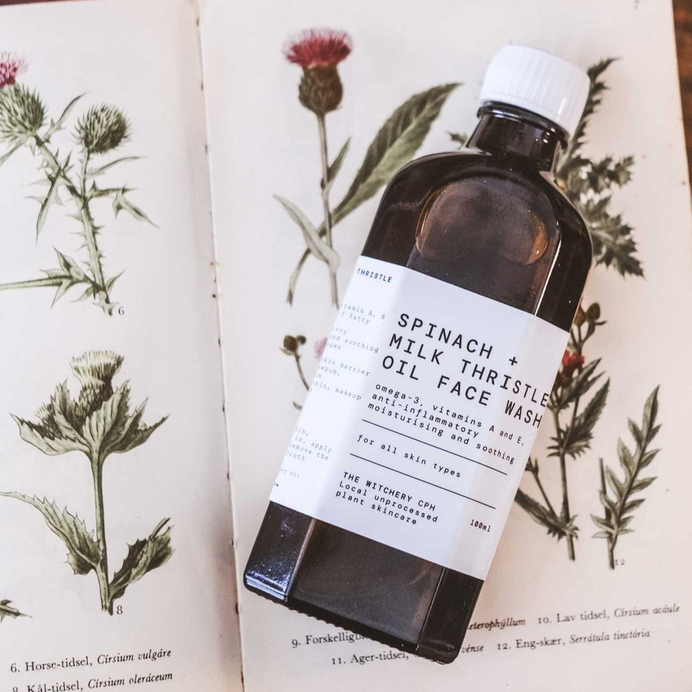An amber glass bottle of Spinach + Milk Thistle Oil Face Wash by The Witchery CPH, featuring a white label and a silver metal cap, set against a plain white background.