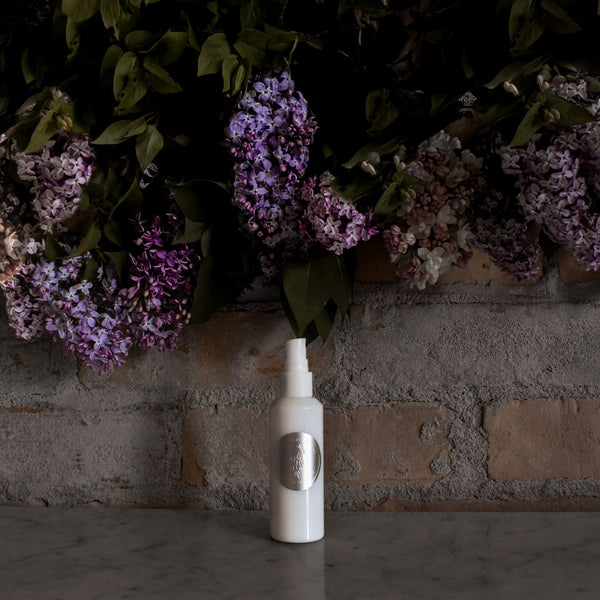 White spray bottle with a logo against a brick wall with purple flowers. TIDELANDS HOUSE Lilac Nectar Artisanal Copper Distillate