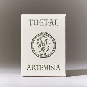 A square cream-colored box standing upright on a light surface against a gray gradient background. The box is printed with the words 'TU·ET·AL' at the top and 'ARTEMISIA' at the bottom in dark olive-green serif lettering, with a central circular illustration of a hand inside a ring.