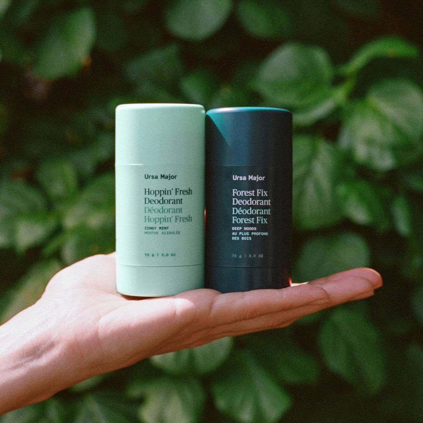 Two deodorant sticks, one light green and one dark blue, held in a hand with a blurred green leafy background.