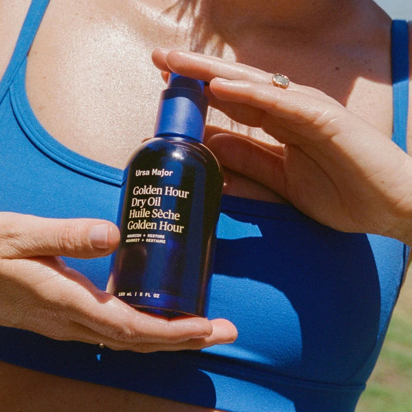 Person holding a bottle of Ursa Major Golden Hour Dry Oil in a natural setting.