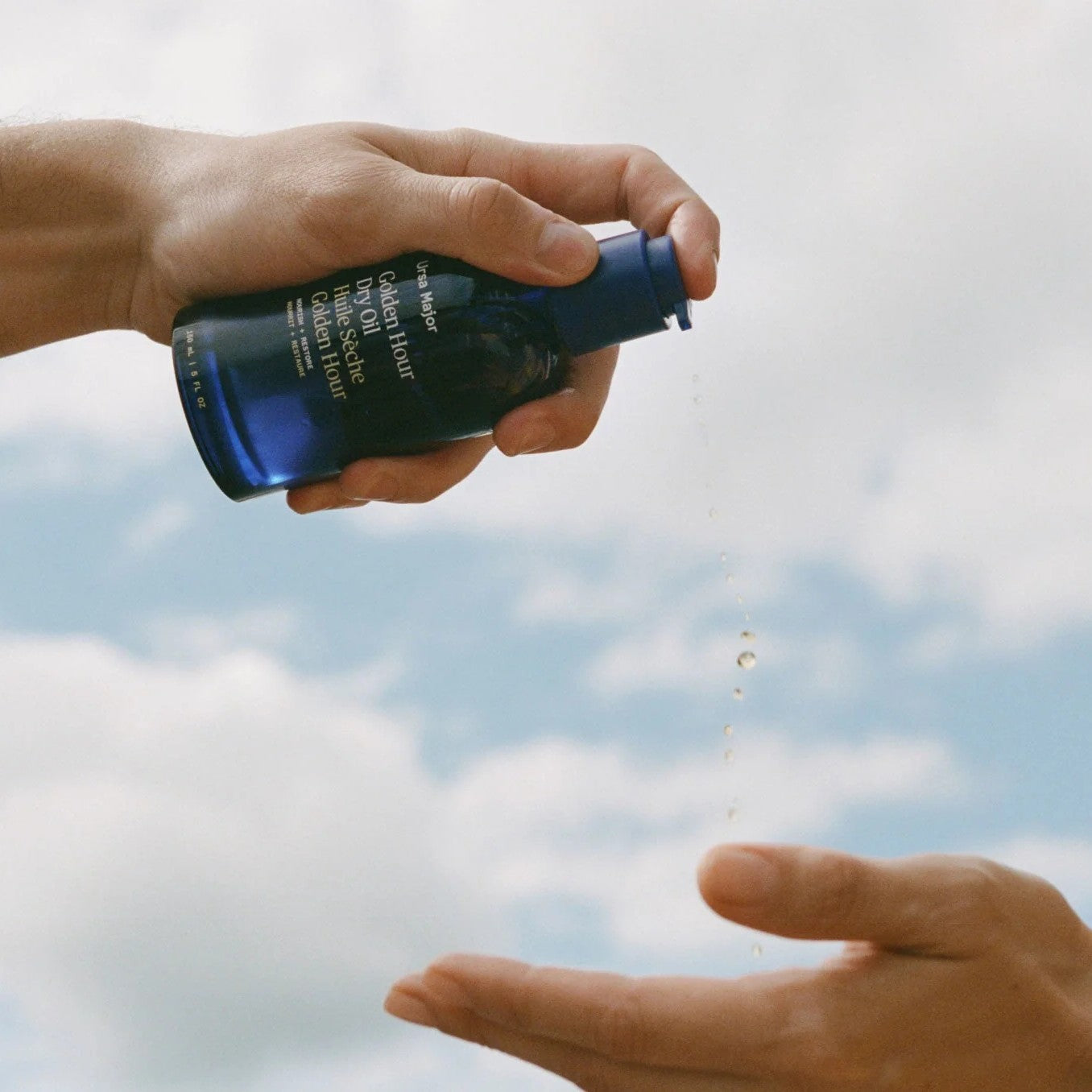 Hand holding a blue bottle with a droplet being released against a cloudy sky. URSA MAJOR Golden Hour Dry Oil