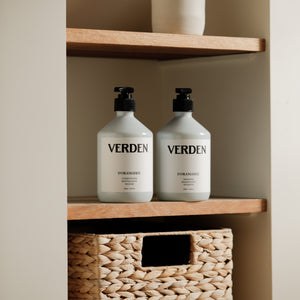 Two white pump bottles labeled VERDEN sit on wooden shelves next to a woven storage basket in a neutral bathroom setting.