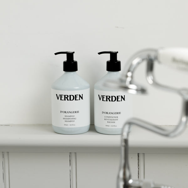 Two white pump bottles of VERDEN haircare products, specifically D'Orangerie Shampoo and Conditioner, sitting on a white shelf next to a blurred chrome faucet.