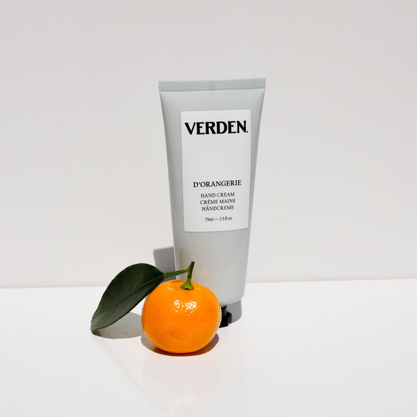 A tube of VERDEN D'ORANGERIE hand cream stands upright on a white surface, with a small orange and a green leaf placed in front of it against a clean white background.