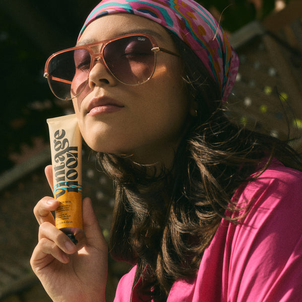 Woman wearing sunglasses and a colorful headscarf, holding a tube of VINTAGE NOON Vacation Skin Overnight Mask and Moisturizer.