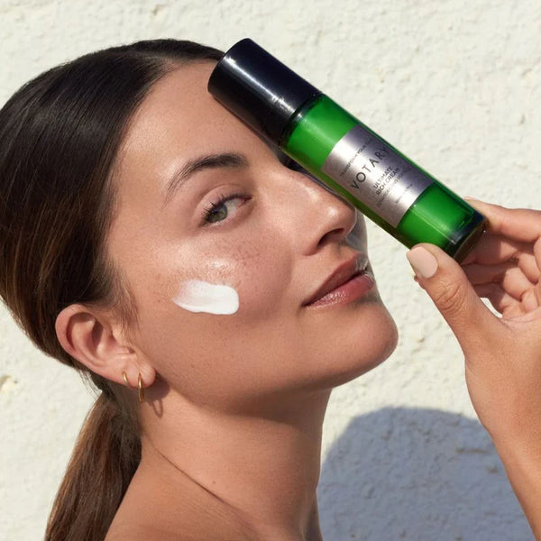 Woman applying a skincare product to her face with a green and black bottle. VOTARY Ultimate Rich Cream