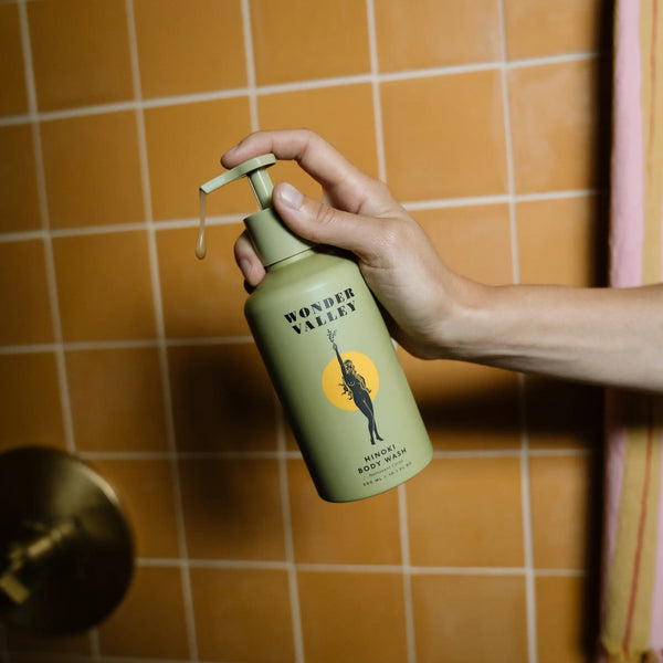 Hand holding a green bottle of Hinoki Body Wash from Wonder Valley against an orange tiled wall.
