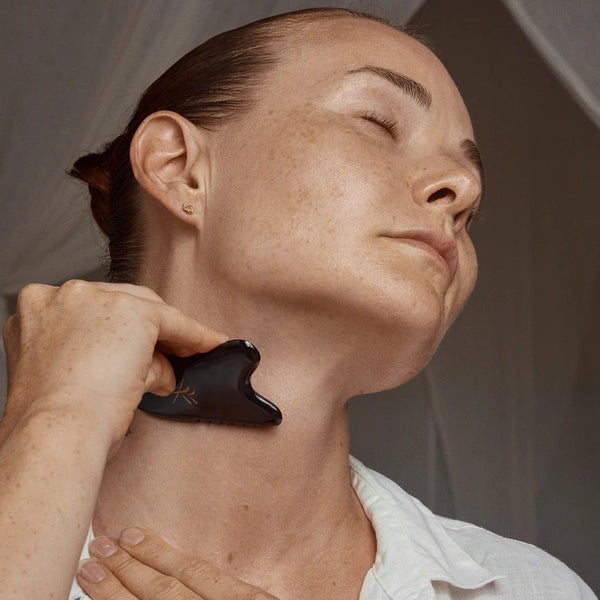 A woman using a black gua sha tool on their neck against a neutral background. YIN YOUR SKIN JALO Advanced Gua Sha Tool