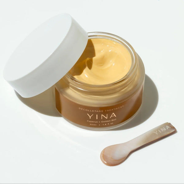 An open jar of YINA Cyprinus + Golden Bell decolletage cream showing a pale yellow cream inside, with its white lid leaning beside it and a small spatula placed in front on a clean white background.